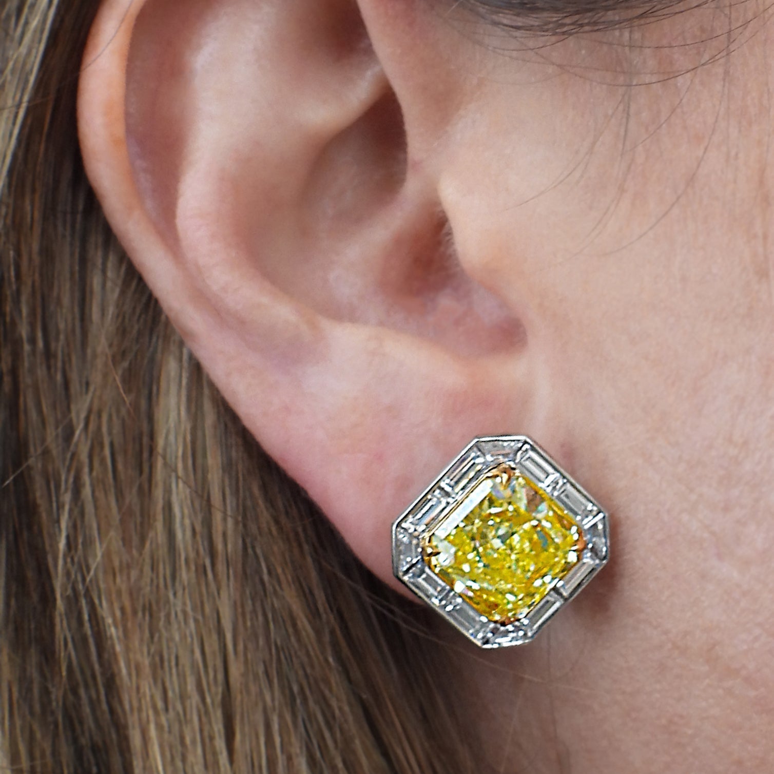 Close-up of a woman wearing 10ct GIA Certified Fancy Intense Yellow Diamond Stud Earrings in Platinum & 18k Gold, highlighting the extravagant yellow diamonds and baguette halo by Vivid Diamonds.
